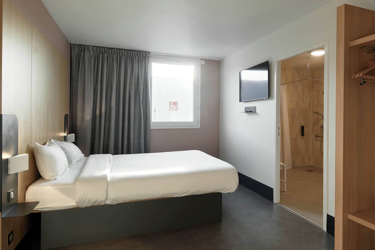 Une chambre d’hôtel moderne équipée d’un lit simple, d’un téléviseur fixé au mur, d’un espace douche et d’une armoire en bois.