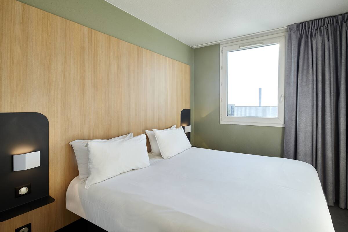 Une chambre d’hôtel avec un grand lit, une tête de lit en bois, deux tables de chevet avec des lampes et une fenêtre avec des rideaux.
