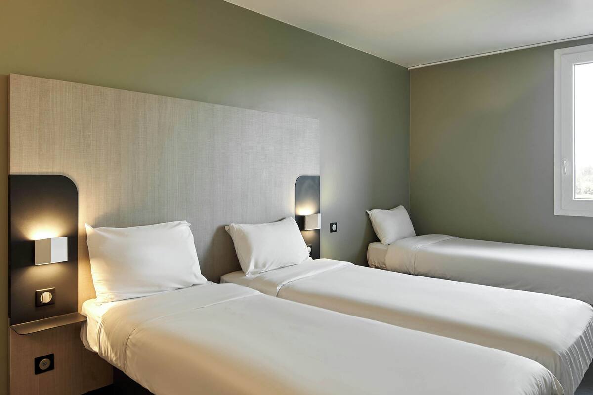Une chambre d’hôtel avec deux lits simples, une table de chevet avec une lampe et une fenêtre laissant entrer la lumière naturelle.