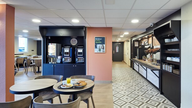 A café area with tables and chairs, a coffee machine, and a wall-mounted sign. - B&B HOTEL Paris Nord Gonesse Parc des Expos (Gonesse)
