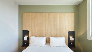 A modern hotel room with a wooden headboard, two bedside tables with illuminated wall sconces, and a neatly made bed. - B&B HOTEL Paris Nord Gonesse Parc des Expos (Gonesse)