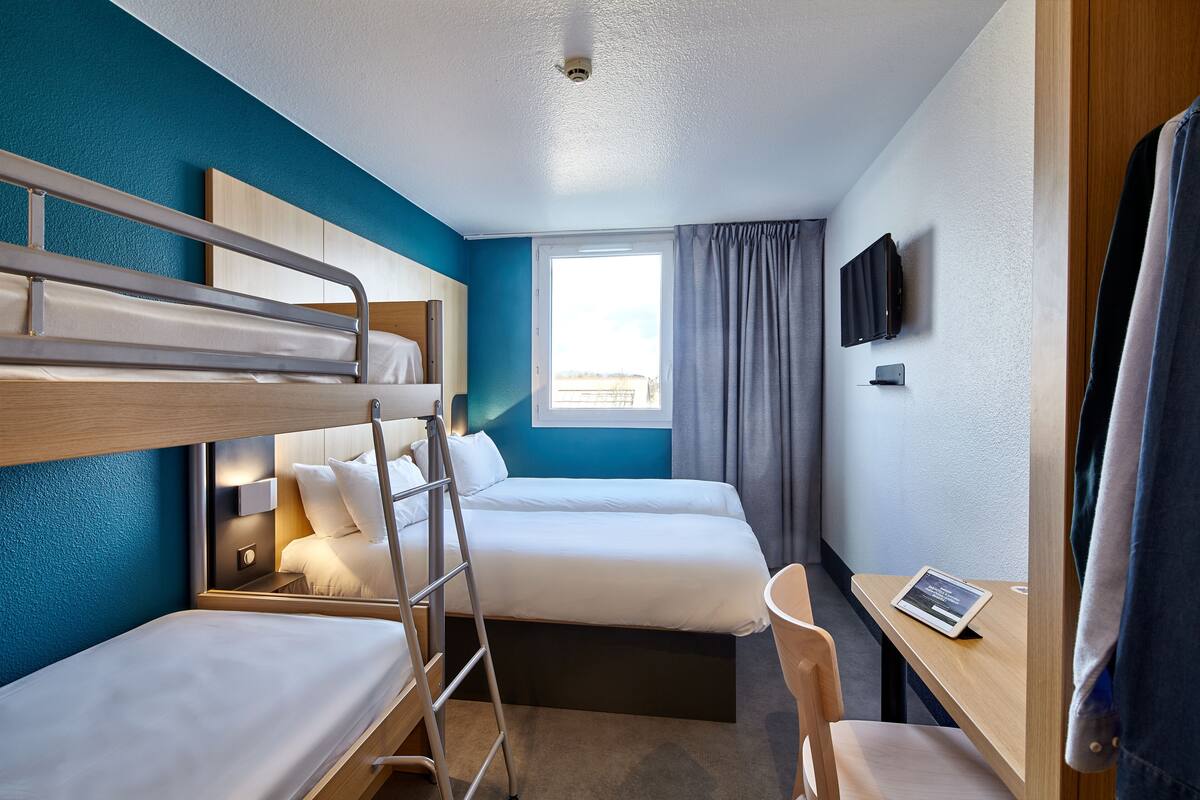 Une chambre d’hôtel avec un lit superposé, un bureau avec un ordinateur portable, une télévision et une fenêtre avec des rideaux.