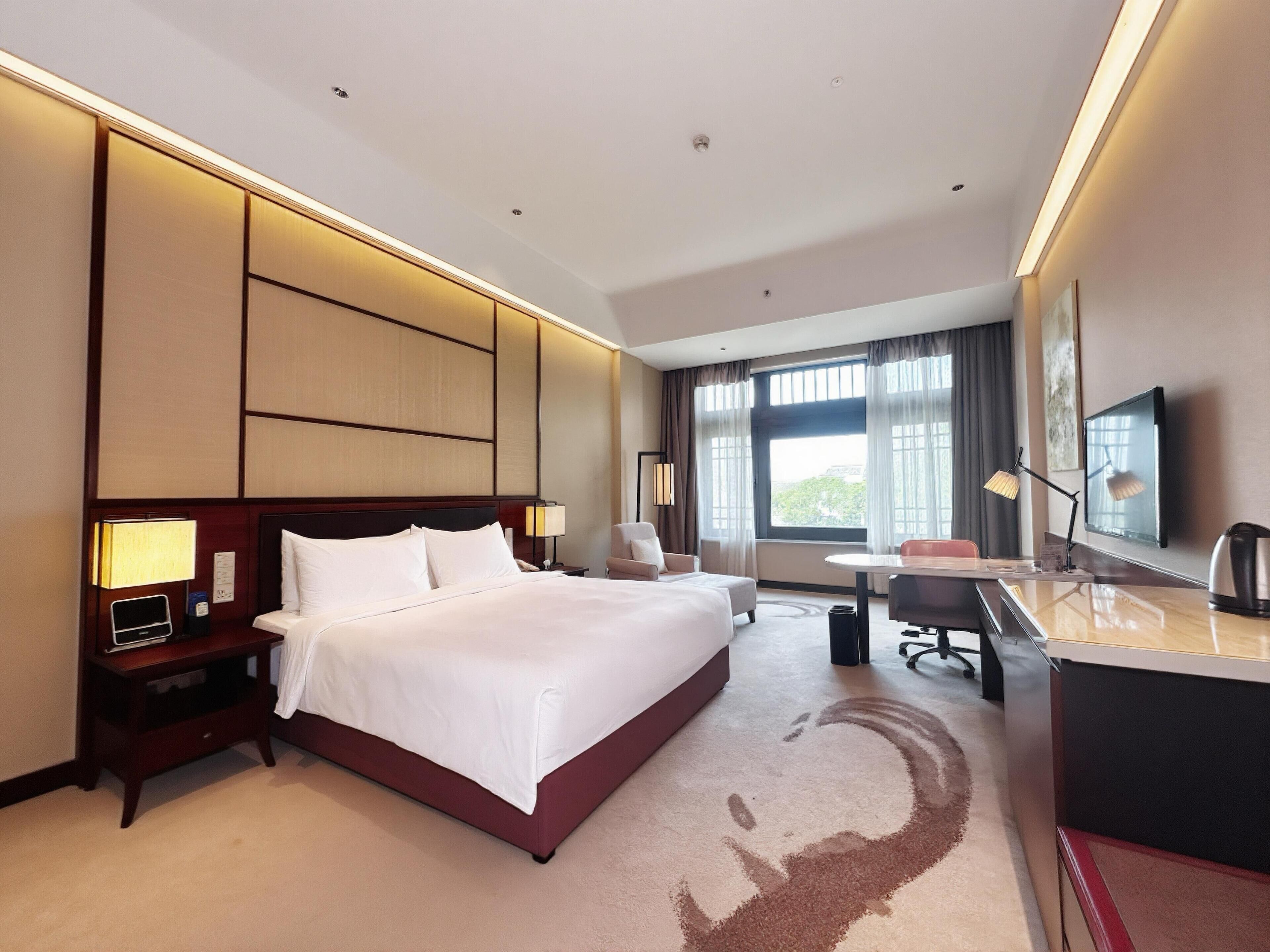 Quarto de hotel com uma cama grande, uma escrivaninha, uma cadeira, uma televisão e uma janela com cortinas.