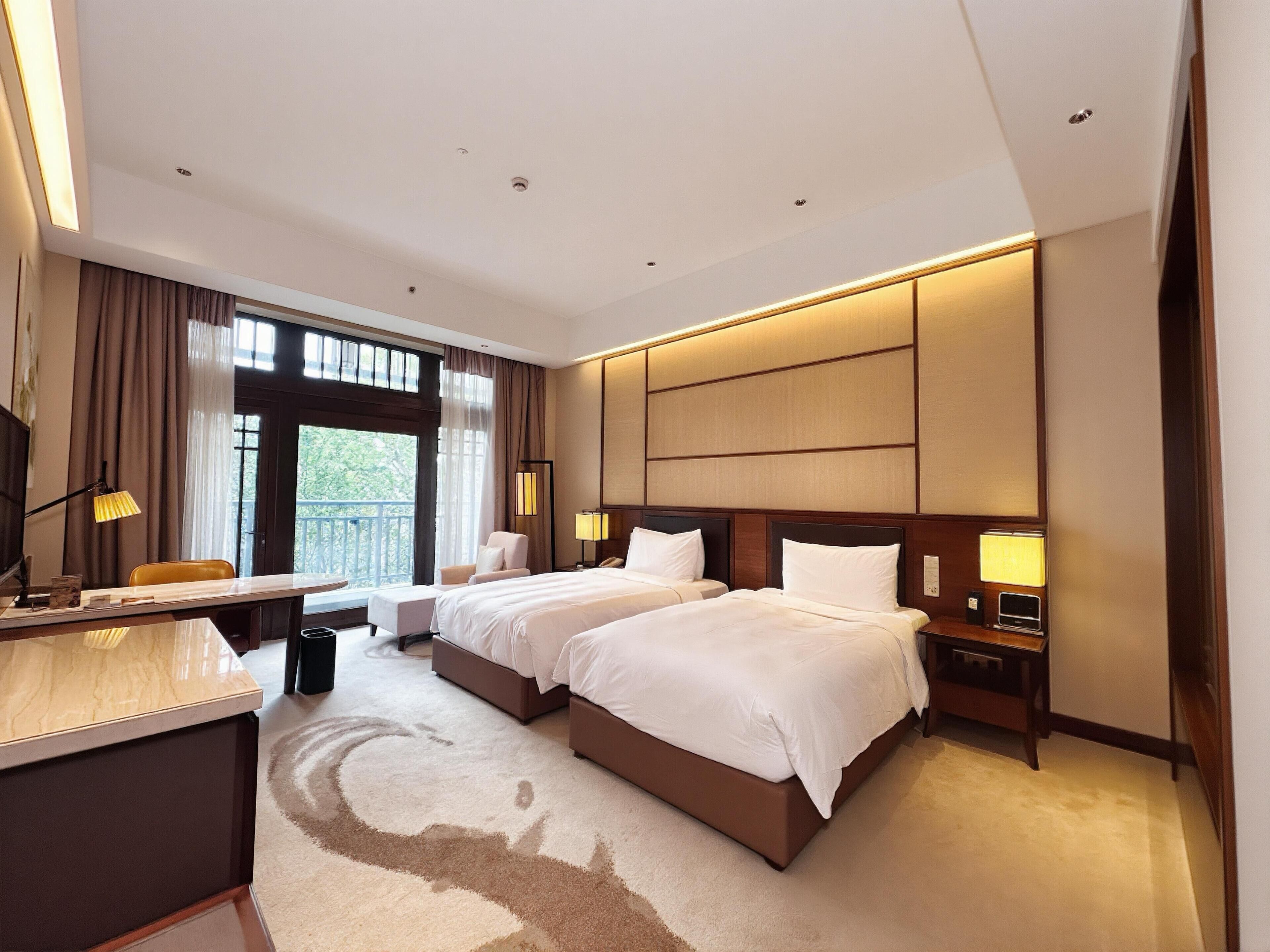 A hotel room with two beds, a desk, a chair, a desk lamp, a nightstand, and a view of the outdoors.