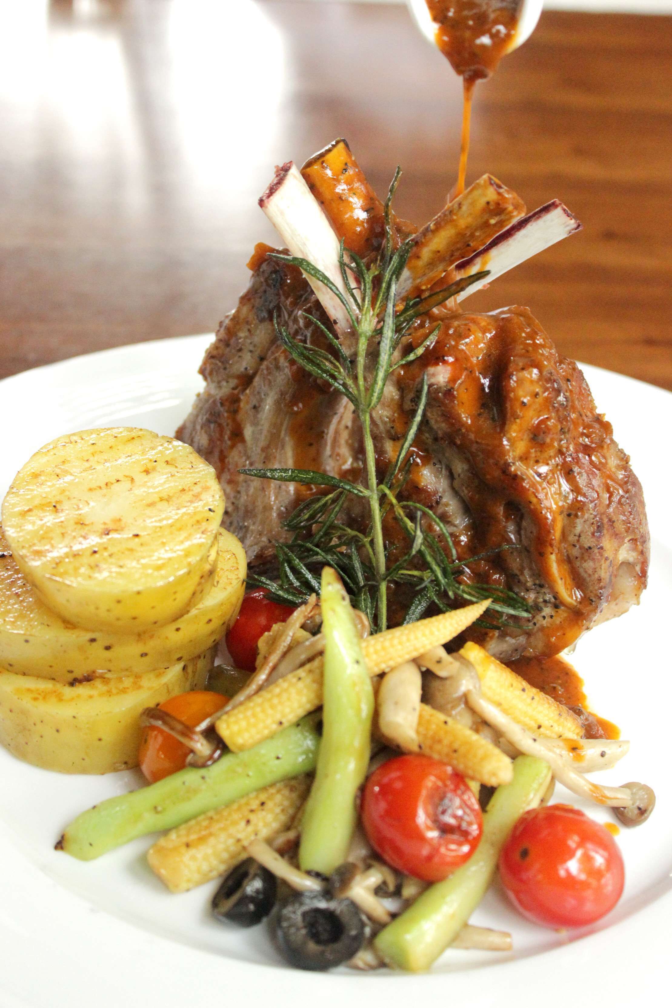 A plate with a roasted lamb shank, roasted potatoes, and a variety of vegetables including carrots, corn, and cherry tomatoes.