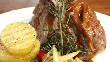 A plate with a roasted lamb shank, roasted potatoes, and a variety of vegetables including carrots, corn, and cherry tomatoes.