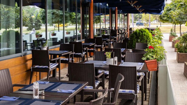 An outdoor dining area with wicker chairs and tables, covered by blue umbrellas, and large windows offering views of the outdoors. - Novotel Sofia (Sofia)