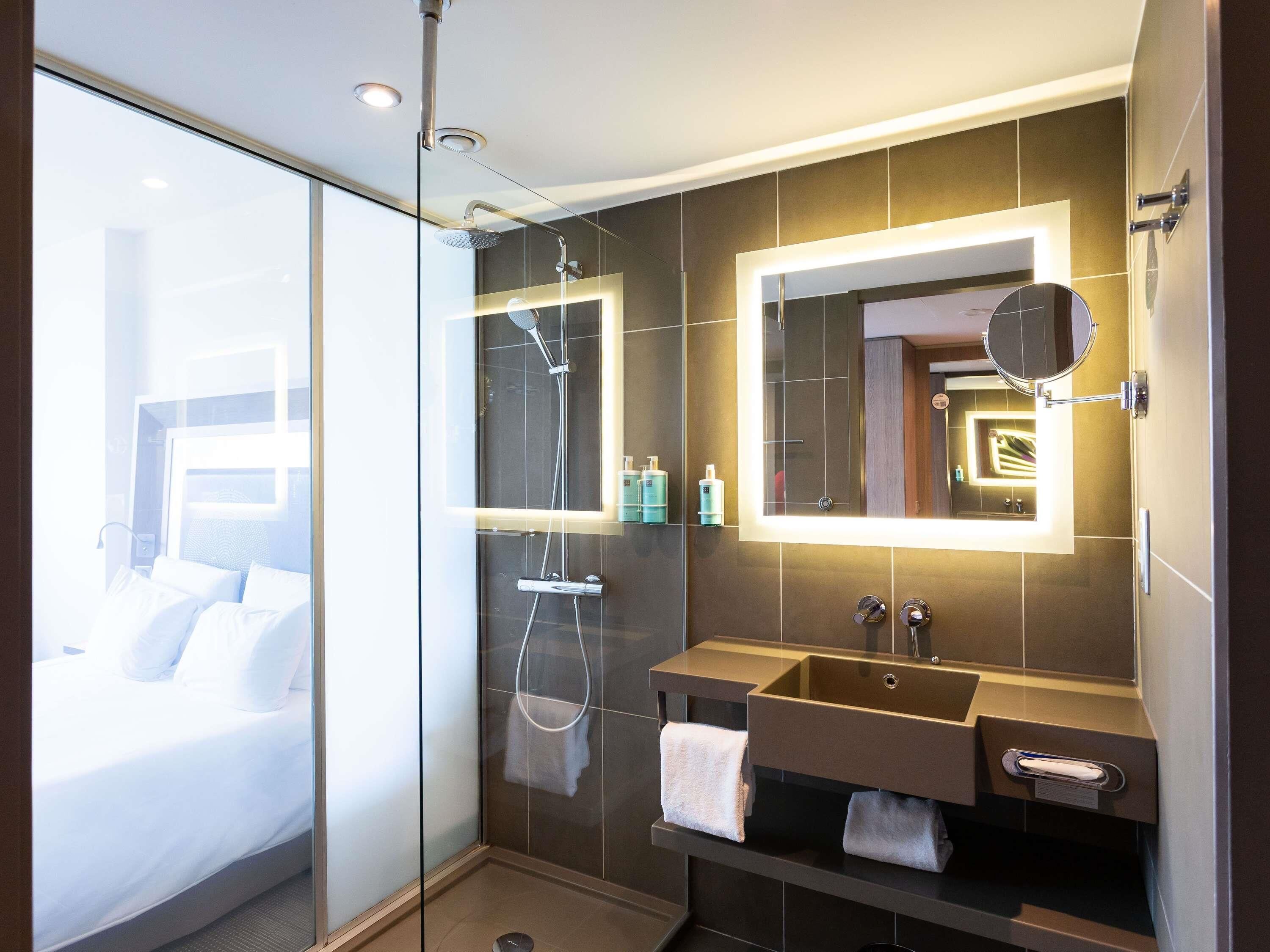 Une salle de bain moderne avec une douche à l’italienne en verre, un meuble-lavabo doté d’un grand miroir et une table de chevet, à travers laquelle on aperçoit le lit.
