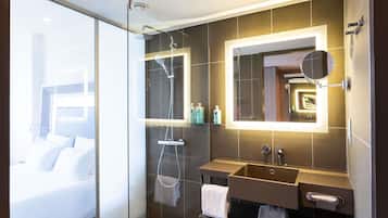Une salle de bain moderne avec une douche à l’italienne en verre, un meuble-lavabo doté d’un grand miroir et une table de chevet, à travers laquelle on aperçoit le lit.
