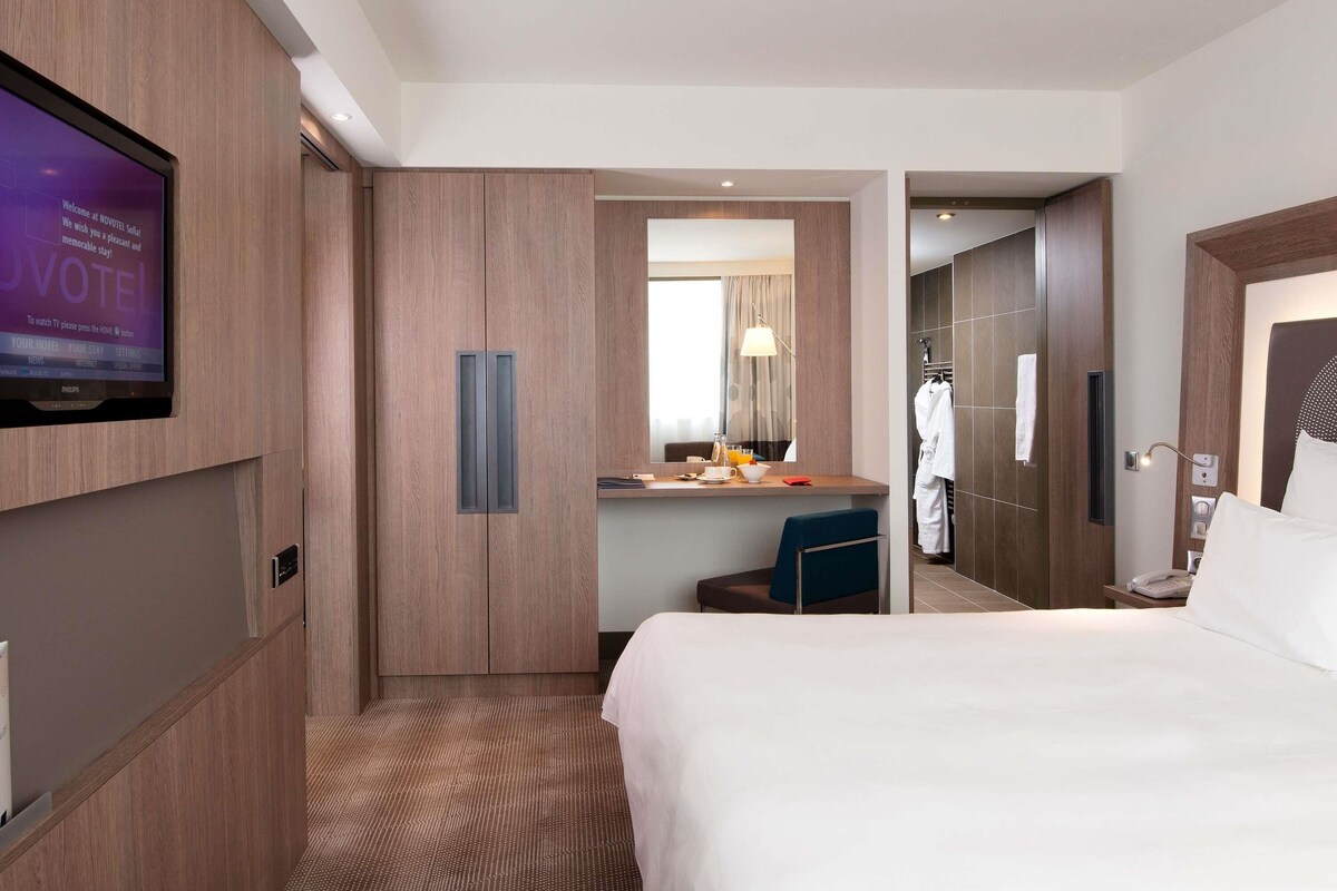 A hotel room with a bed, a desk with a chair, a television mounted on the wall, and a bathroom visible through an open door.