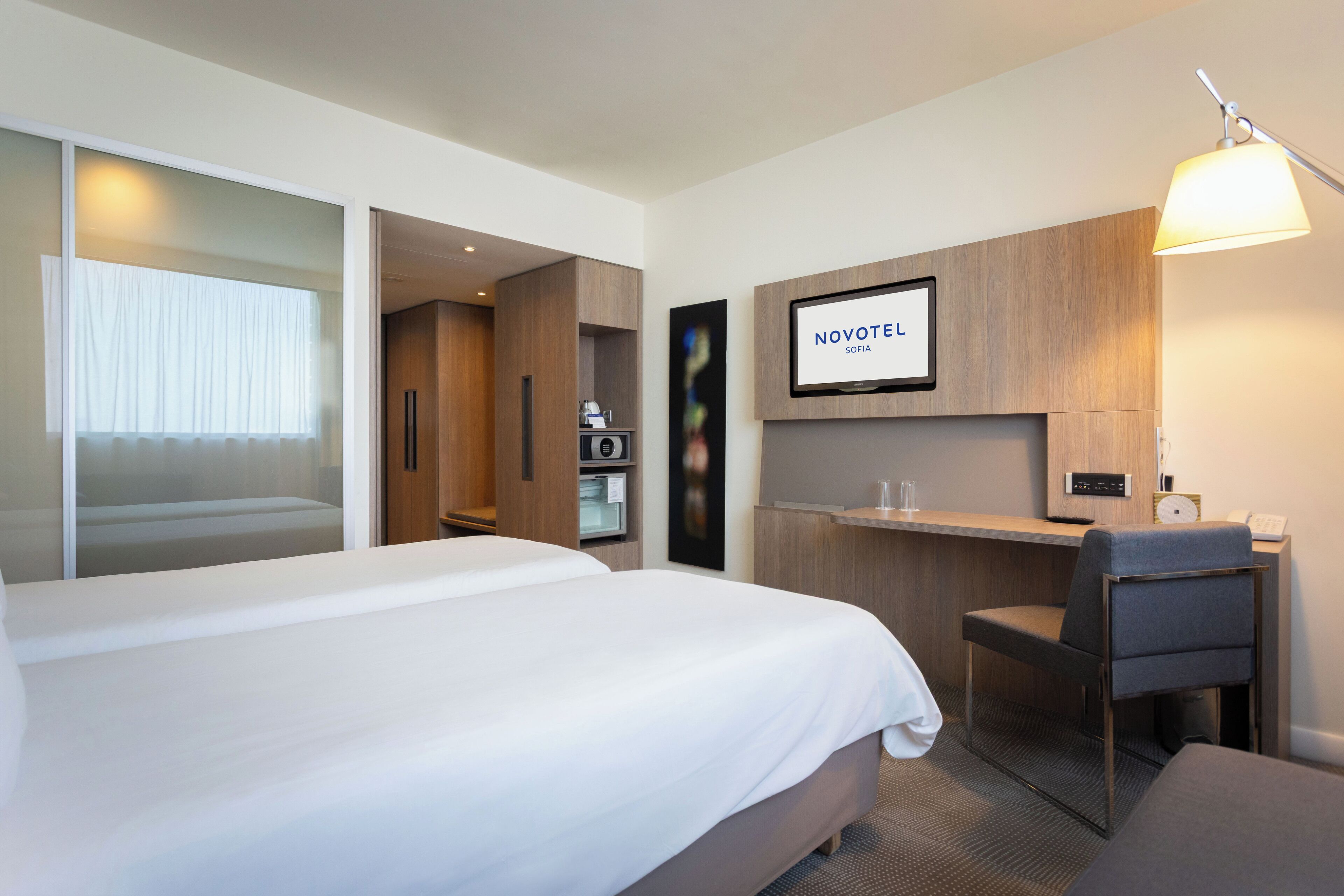 A hotel room with a large bed, a desk, a chair, and a TV screen displaying the Novotel logo.