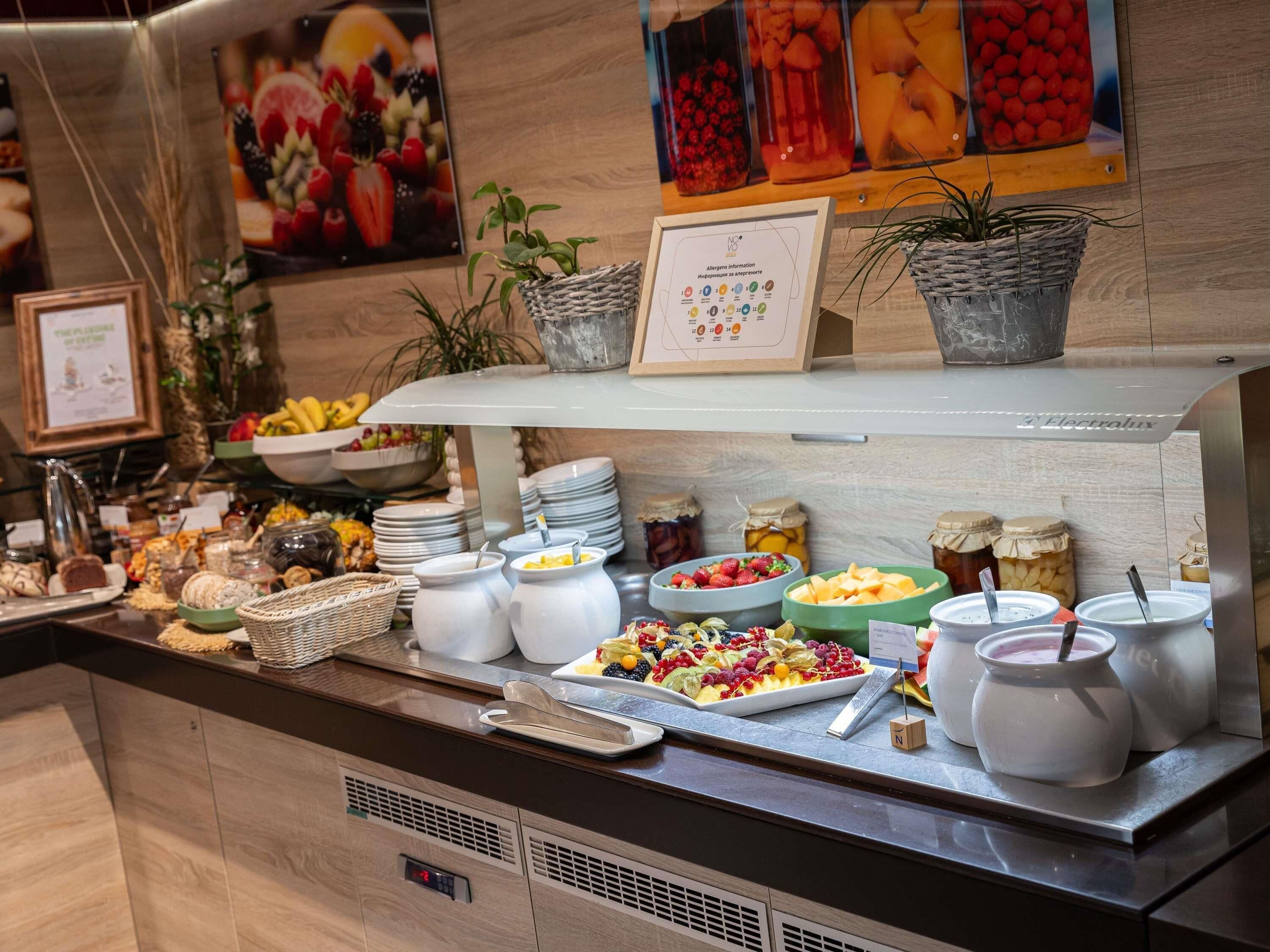Un bufé con diversos alimentos, como frutas, quesos y panes, dispuesto en un mostrador con fondo de madera y plantas decorativas.
