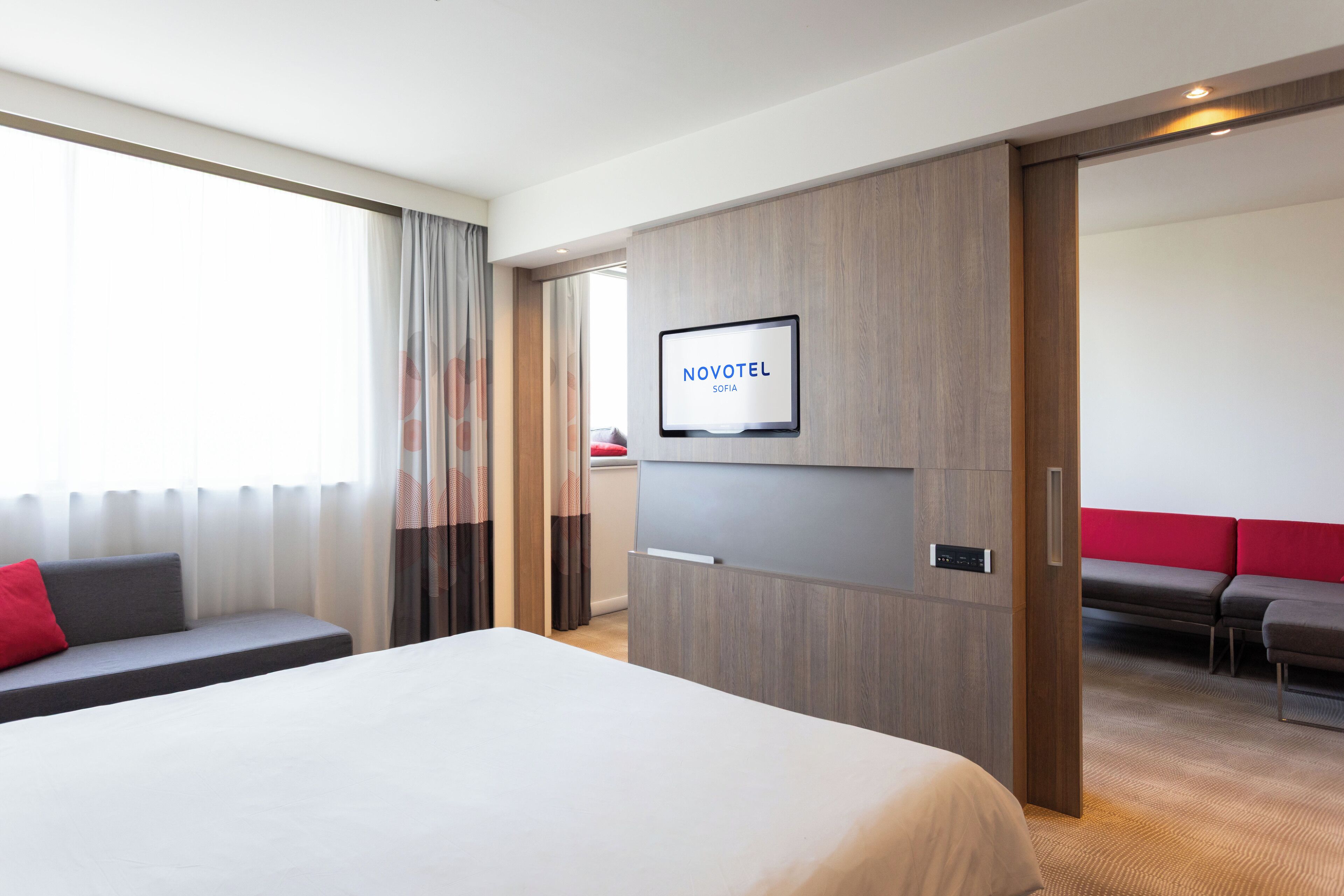 Habitación de hotel con una cama, un televisor fijado en la pared, una zona de estar con un cojín rojo y una pared de paneles de madera.