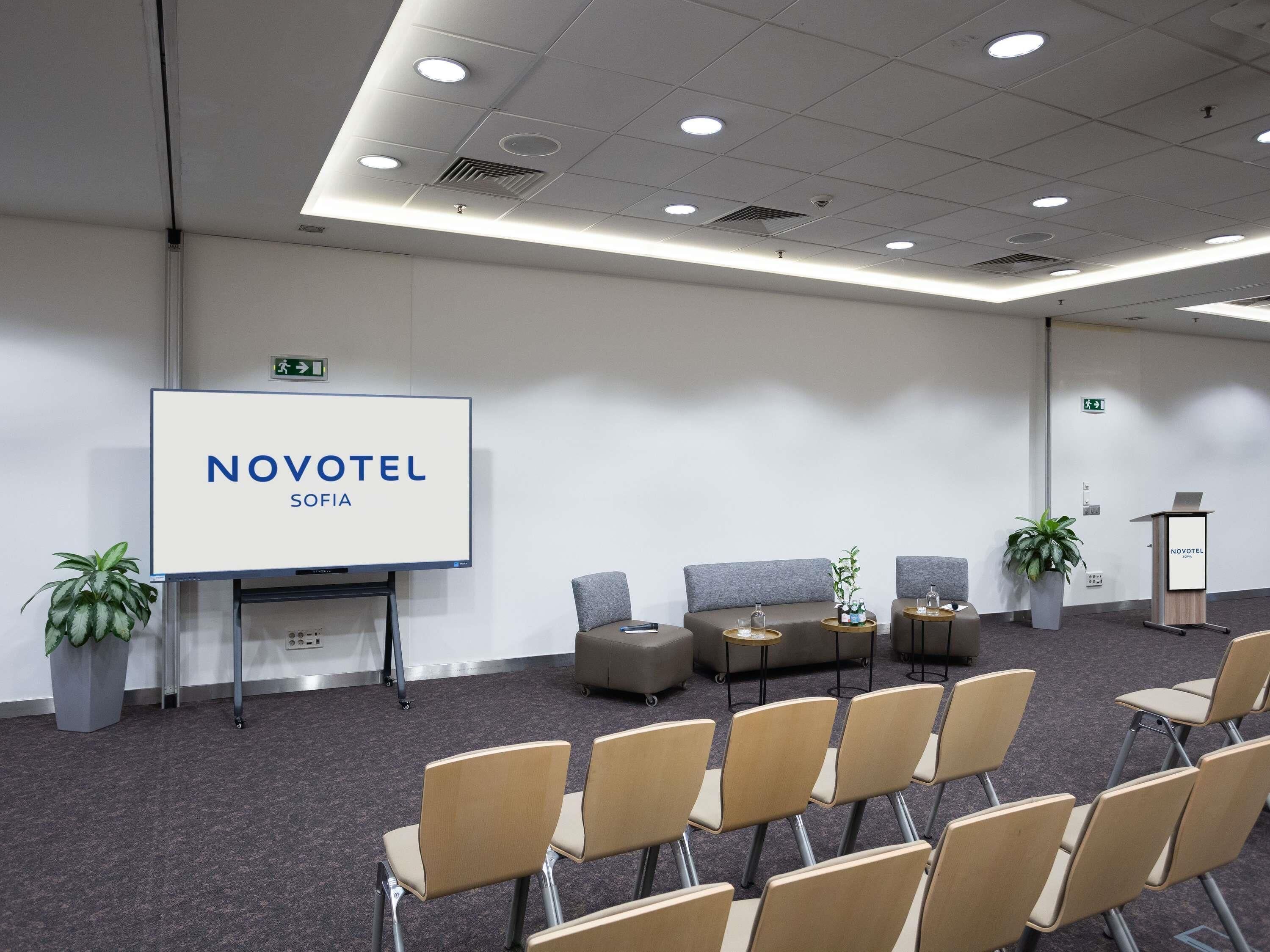 A conference room with a large screen displaying 'NOVOTEL SOFIA,' a sofa, chairs, a small table, and potted plants.
