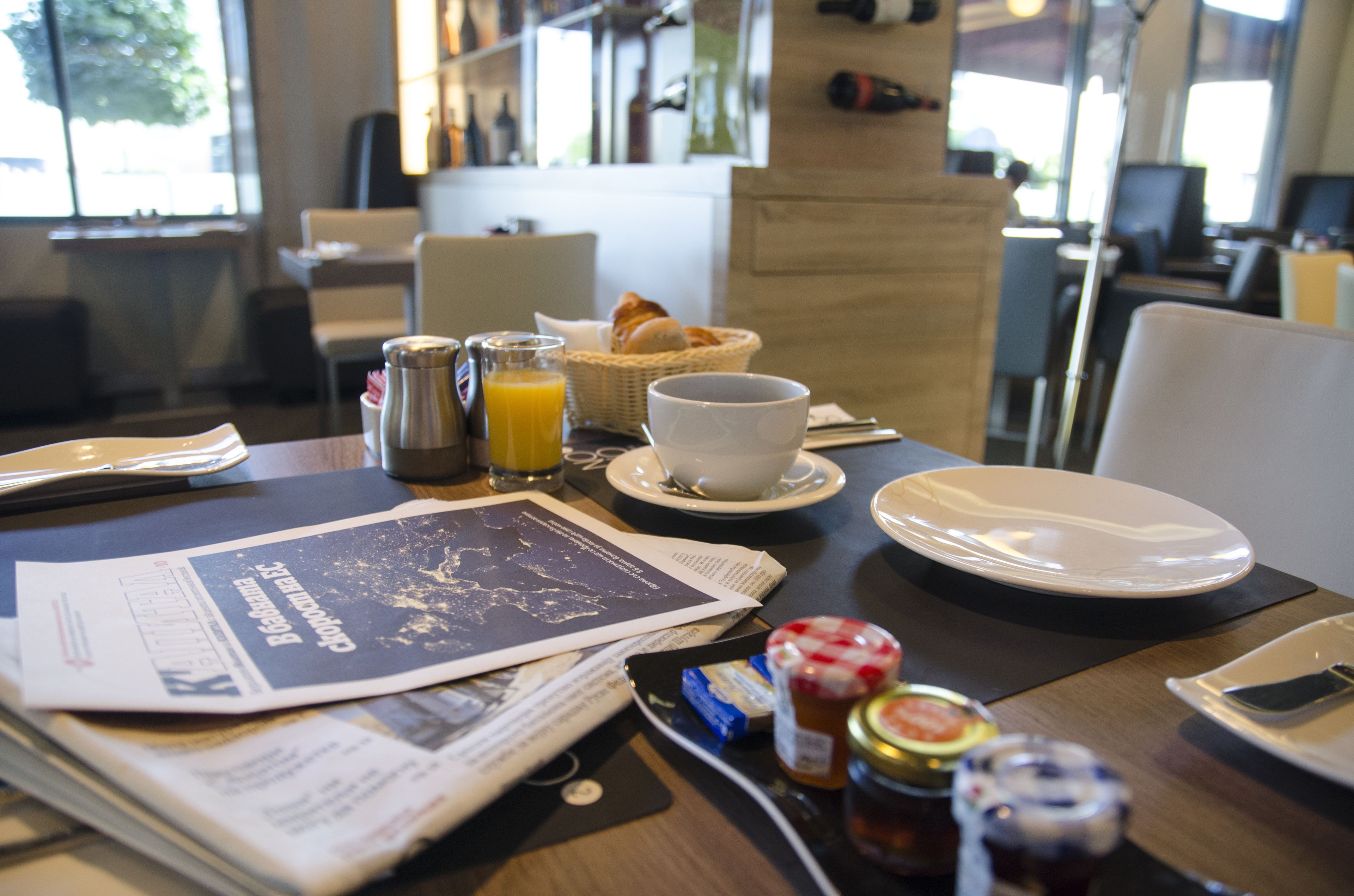 A breakfast table with a newspaper, a basket of croissants, a glass of orange juice, and various condiments.