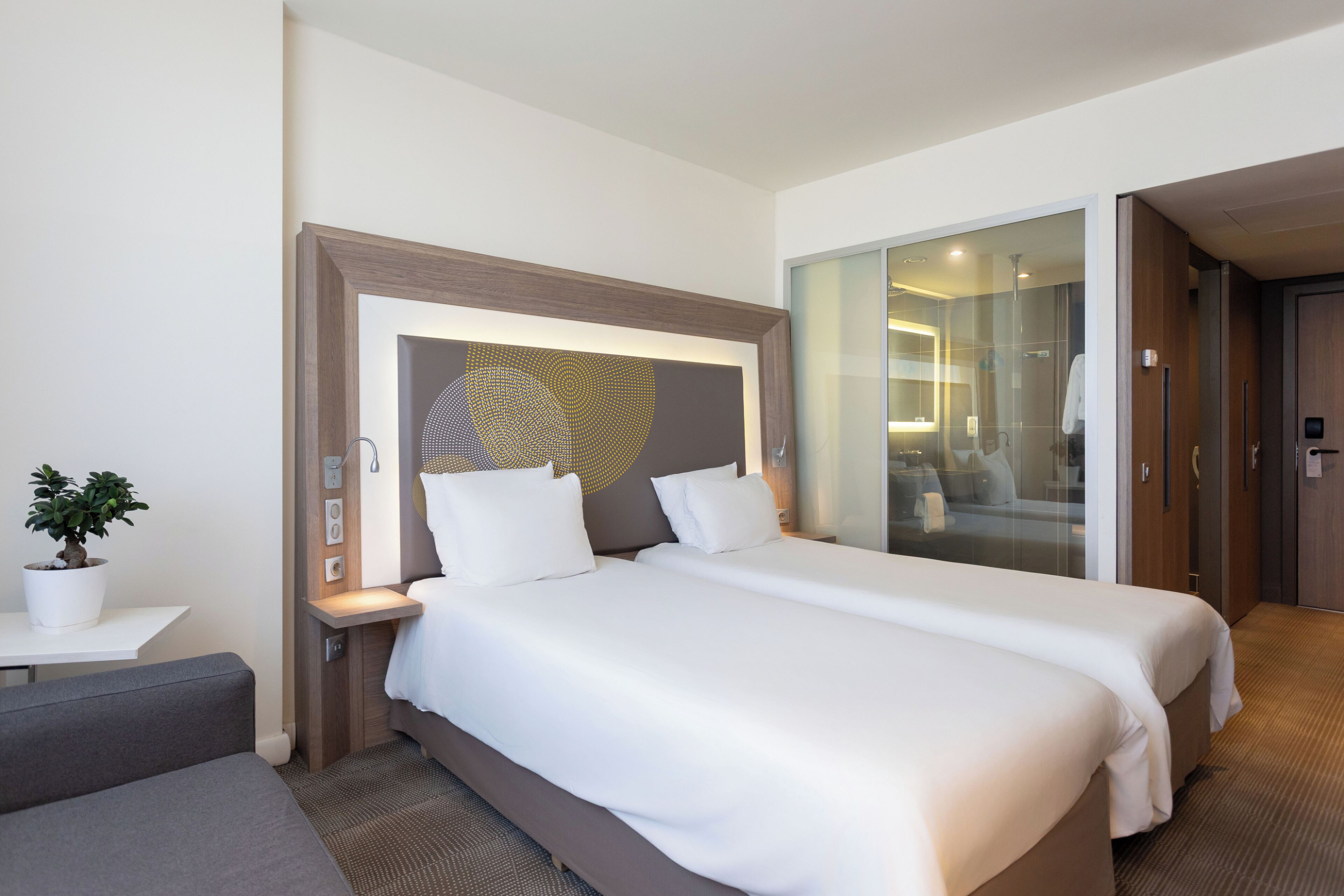 A modern hotel room with two beds, a small table with a potted plant, and a glass shower area.