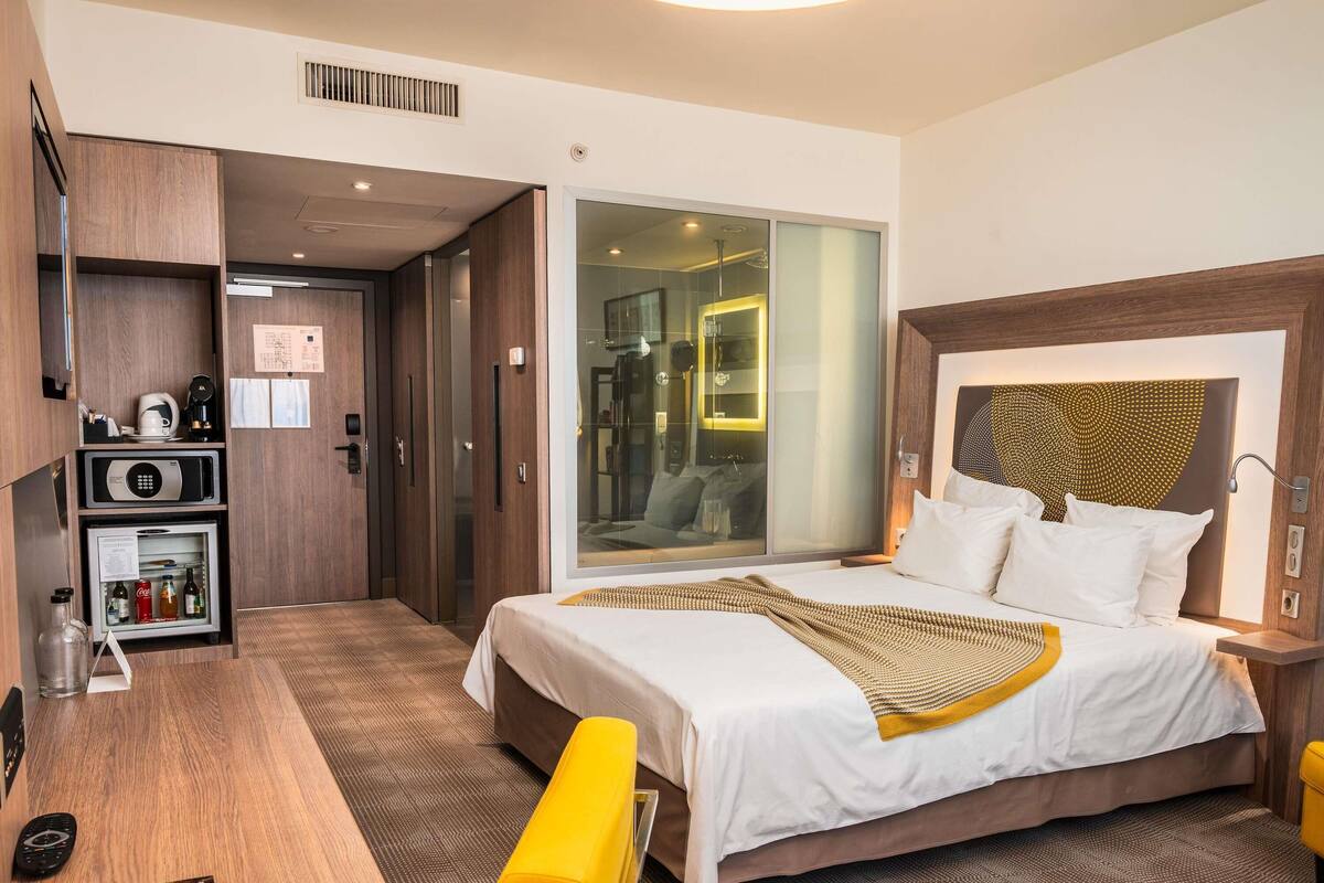 A hotel room with a bed, bedside table, a desk with a microwave, a refrigerator, and a bench.