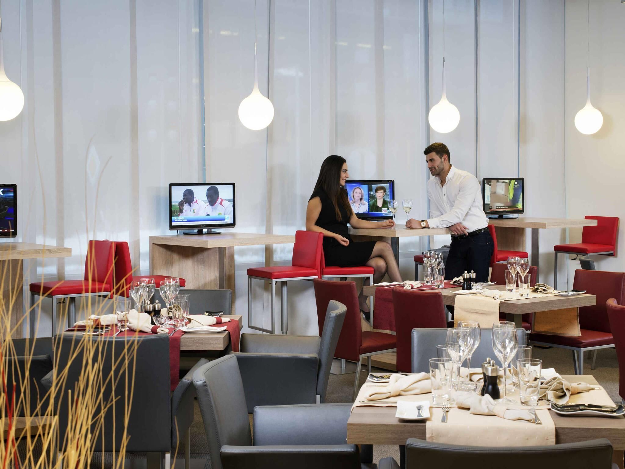 A modern restaurant with red and grey chairs, tables set for dining, and multiple screens displaying content.