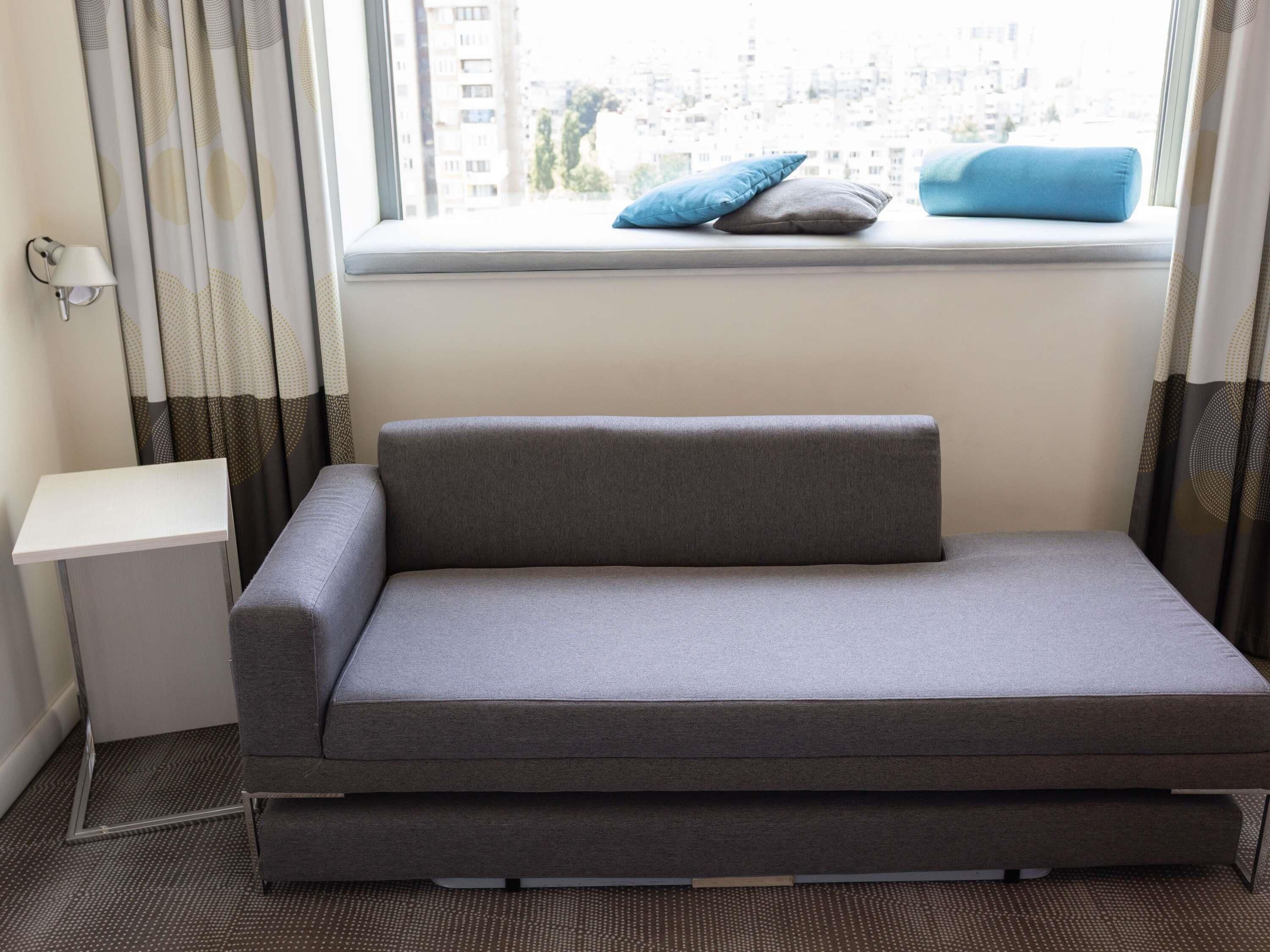 A grey sofa with a cushion, a small white side table, and a window with curtains.