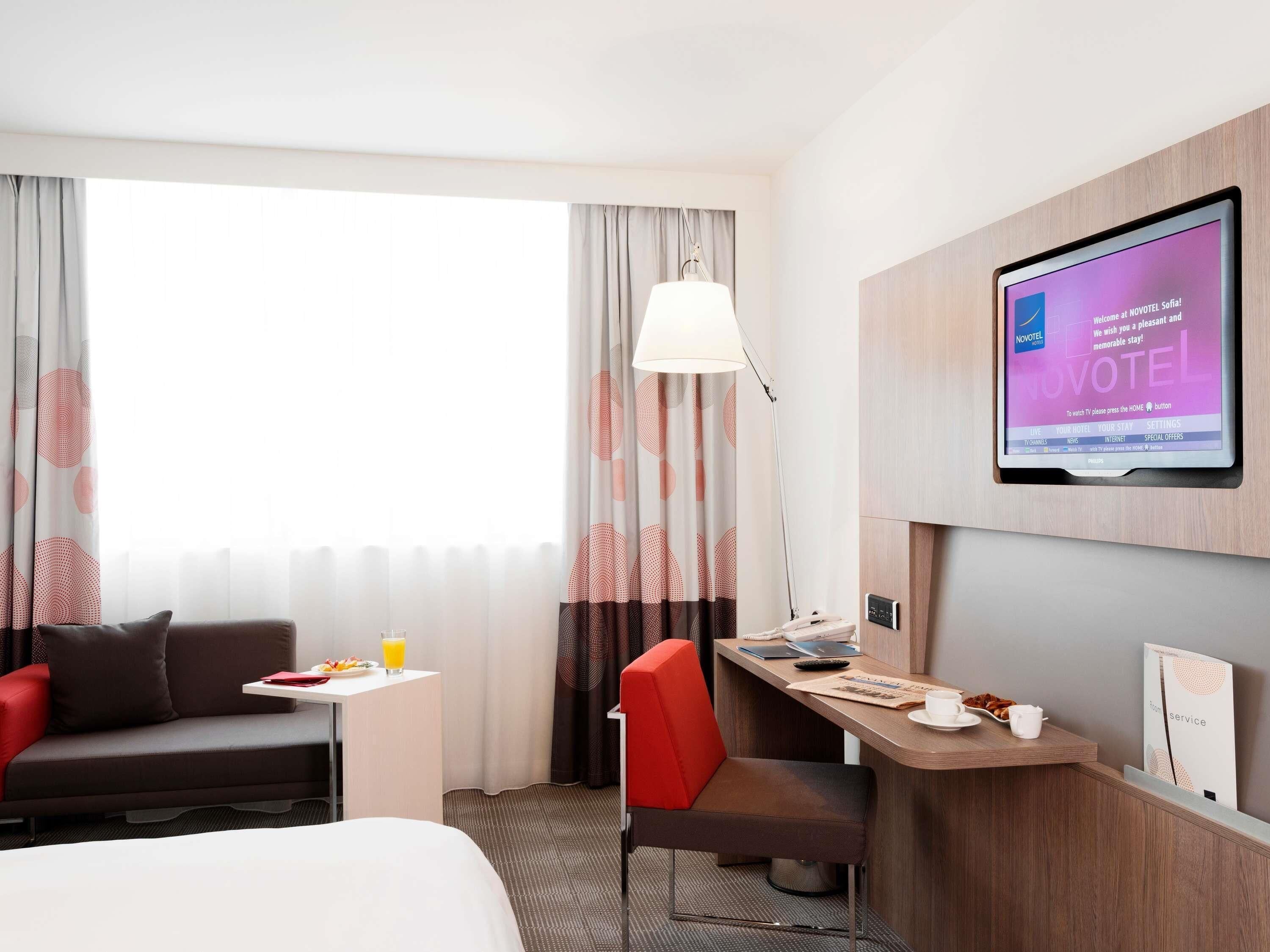 A hotel room with a bed, a desk with a chair, a TV mounted on the wall, and a window with curtains.