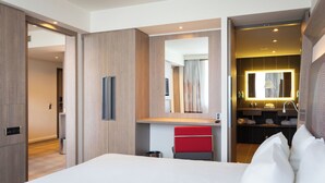 A hotel room with a large bed, a red chair, a vanity with a mirror, and a small bathroom. - Novotel Sofia (Sofia)