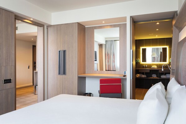 Une chambre d’hôtel avec un grand lit, un fauteuil rouge, une coiffeuse avec un miroir et une petite salle de bain.