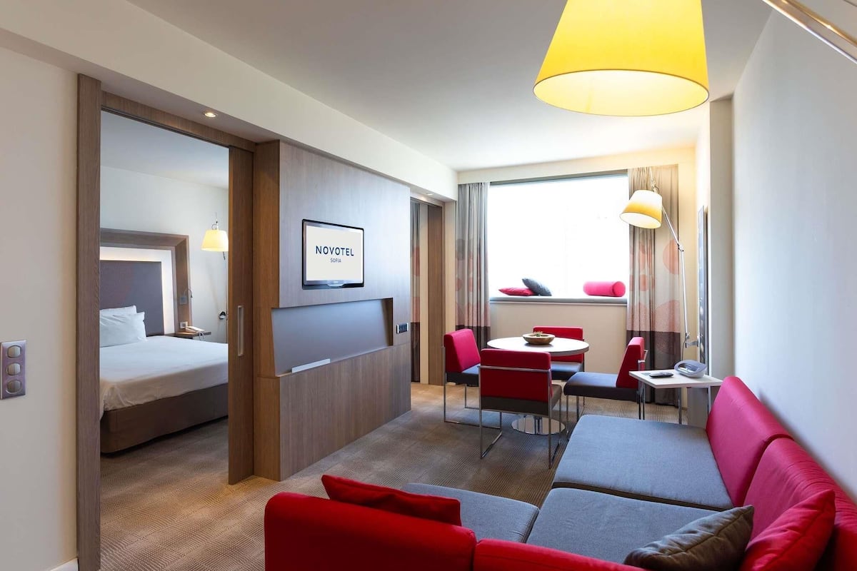 Habitación de hotel con cama, sofá, zona de comedor con sillas, televisión y ventana con cortinas.