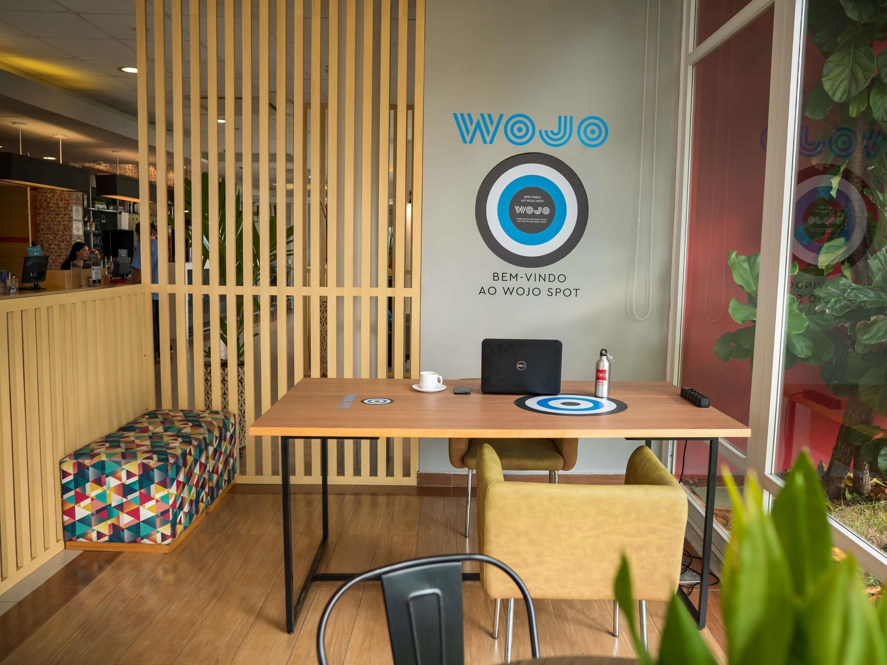 Een moderne kantoorruimte met een houten scheidingswand, een bureau met een laptop, een witte beker en een blikje drinken. Op de voorgrond staat een kleurrijke bank en een plant.