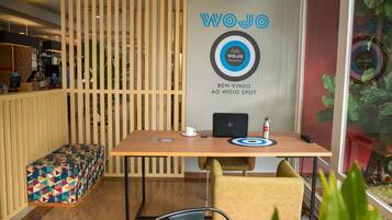 Een moderne kantoorruimte met een houten scheidingswand, een bureau met een laptop, een witte beker en een blikje drinken. Op de voorgrond staat een kleurrijke bank en een plant.