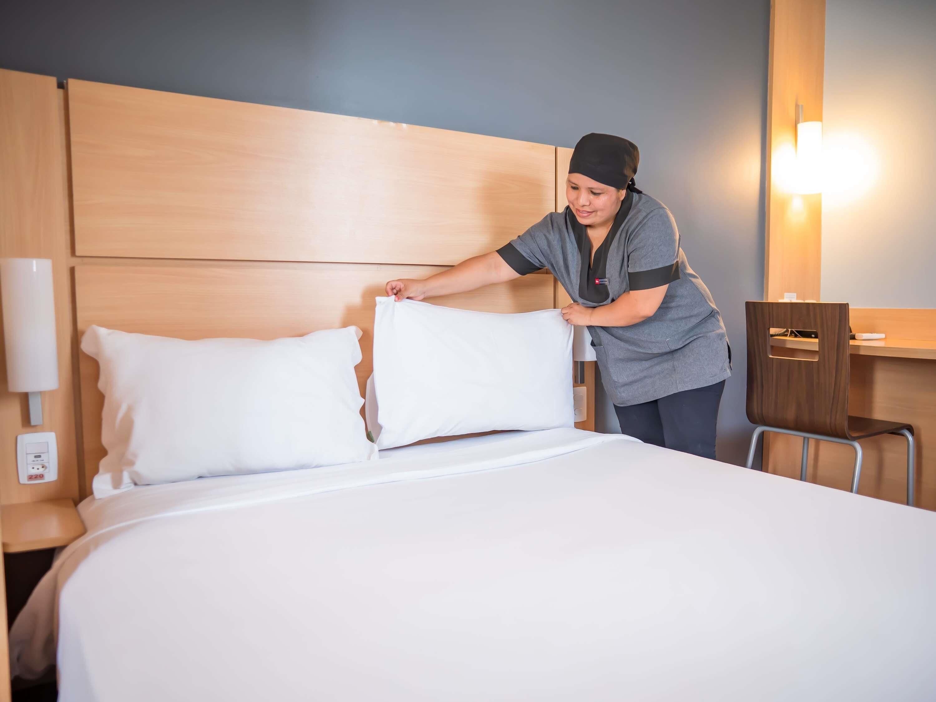 Una habitación de hotel con cabecera de madera, cama con ropa blanca, mesita de noche con lámpara y escritorio con silla.