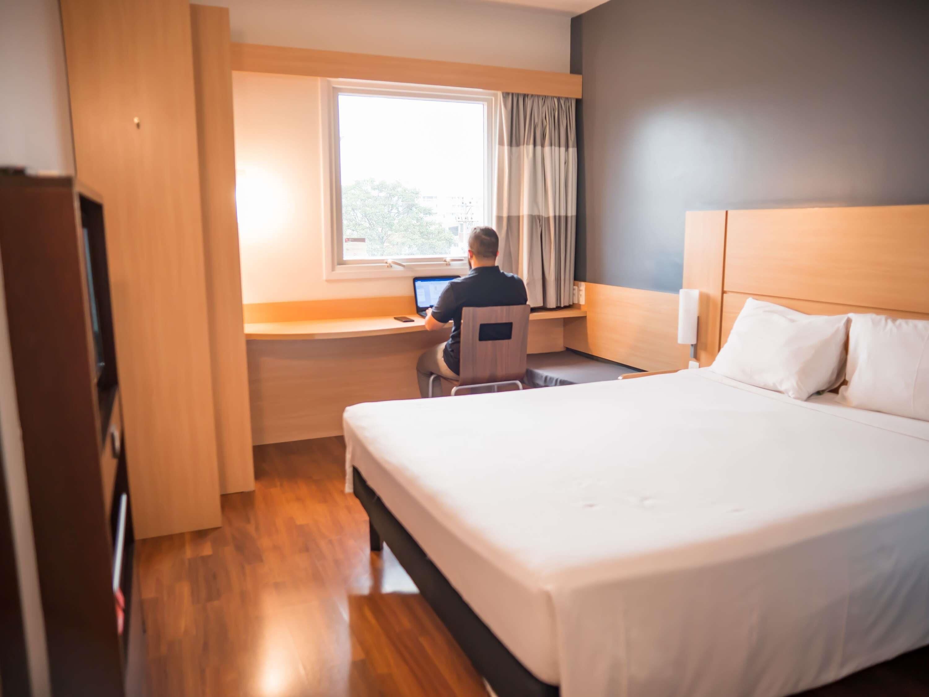Una habitación de hotel con cama, escritorio con computadora portátil, ventana con cortinas y piso de madera.