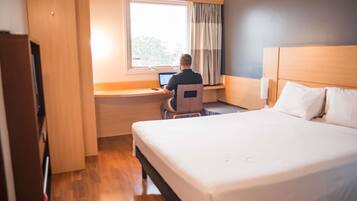 Una habitación de hotel con cama, escritorio con computadora portátil, ventana con cortinas y piso de madera.