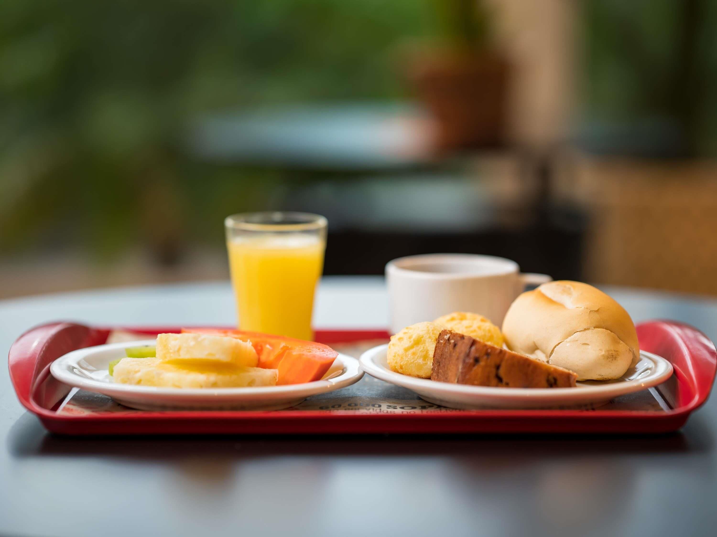Bandeja de desayuno con vaso de jugo de naranja, un tazón de sopa y dos productos de pastelería sobre una mesa con fondo desenfocado.