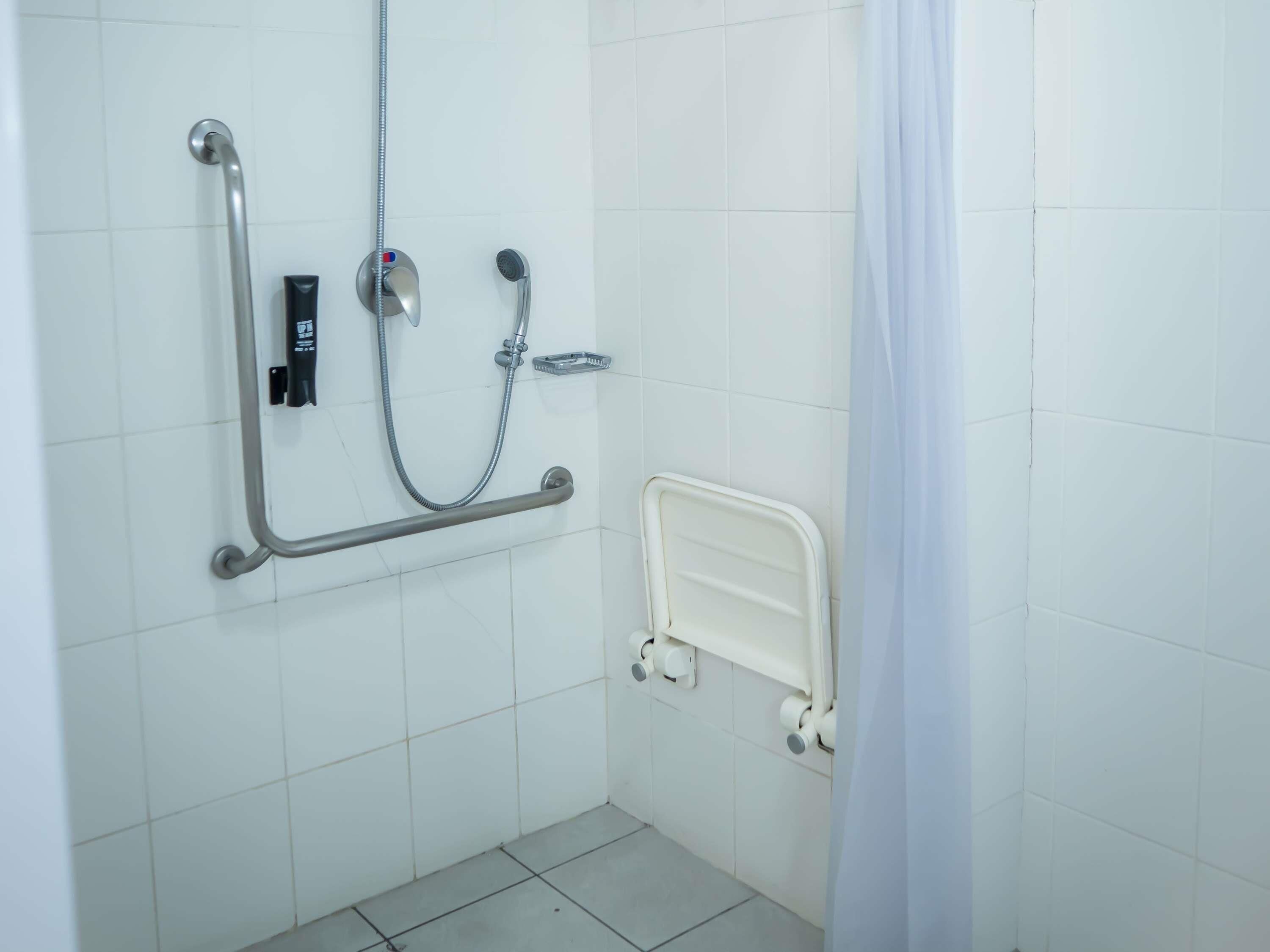 A bathroom with white tiled walls, a shower with a handheld showerhead, a grab bar, and a white toilet.