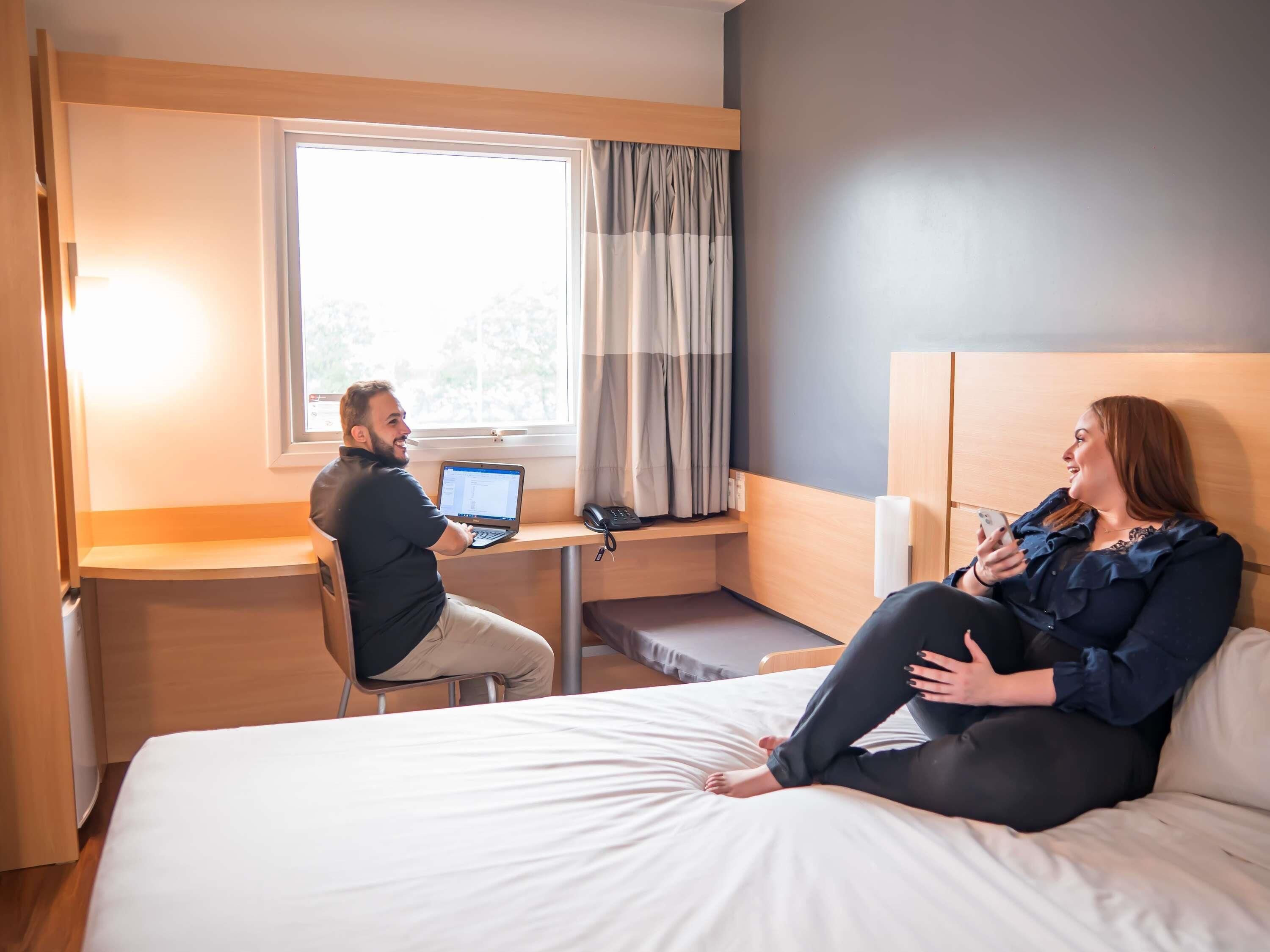 Een hotelkamer met een bed, een bureau met een laptop, een nachtkastje en een raam met gordijnen.
