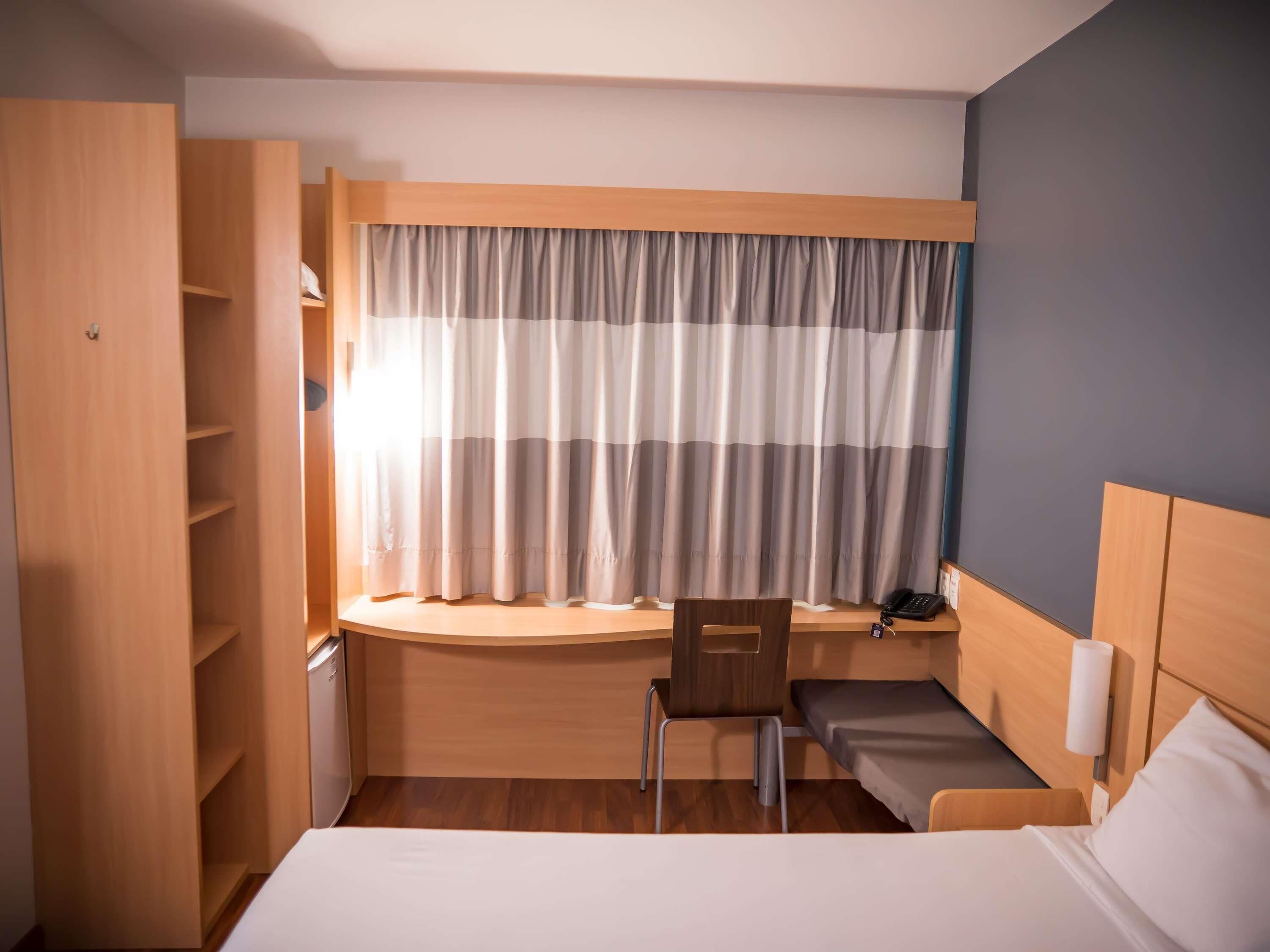 A compact hotel room with a bed, a desk, a chair, and a small seating area.