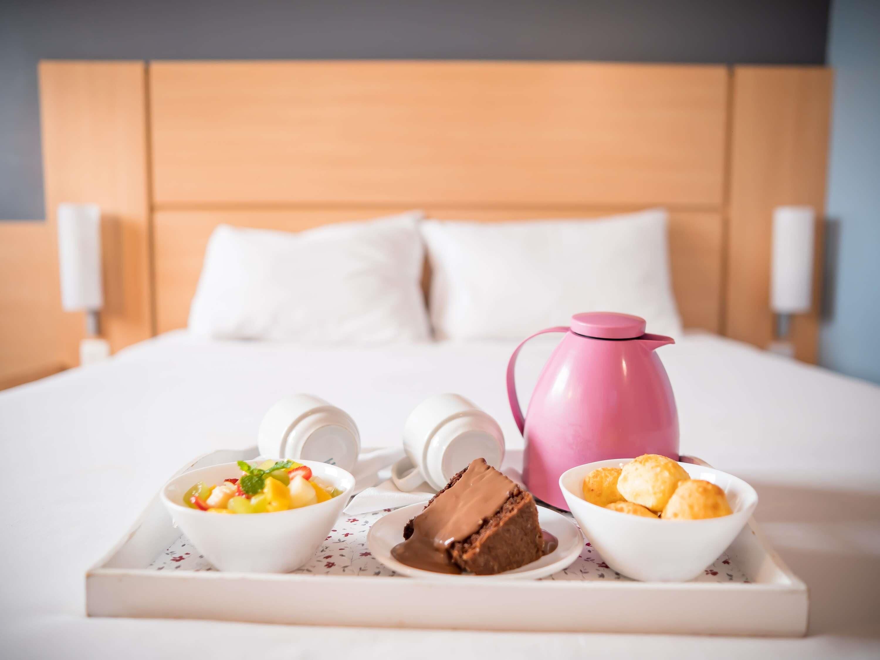 A neatly made breakfast tray with a pink coffee pot, a bowl of fruit salad, a chocolate dessert, and a bowl of pastries on a hotel bed.