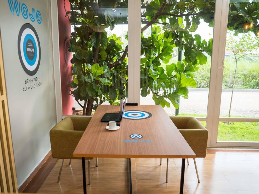 A conference room with a wooden table, two chairs, a laptop, a cup, and a large window with a view of greenery.