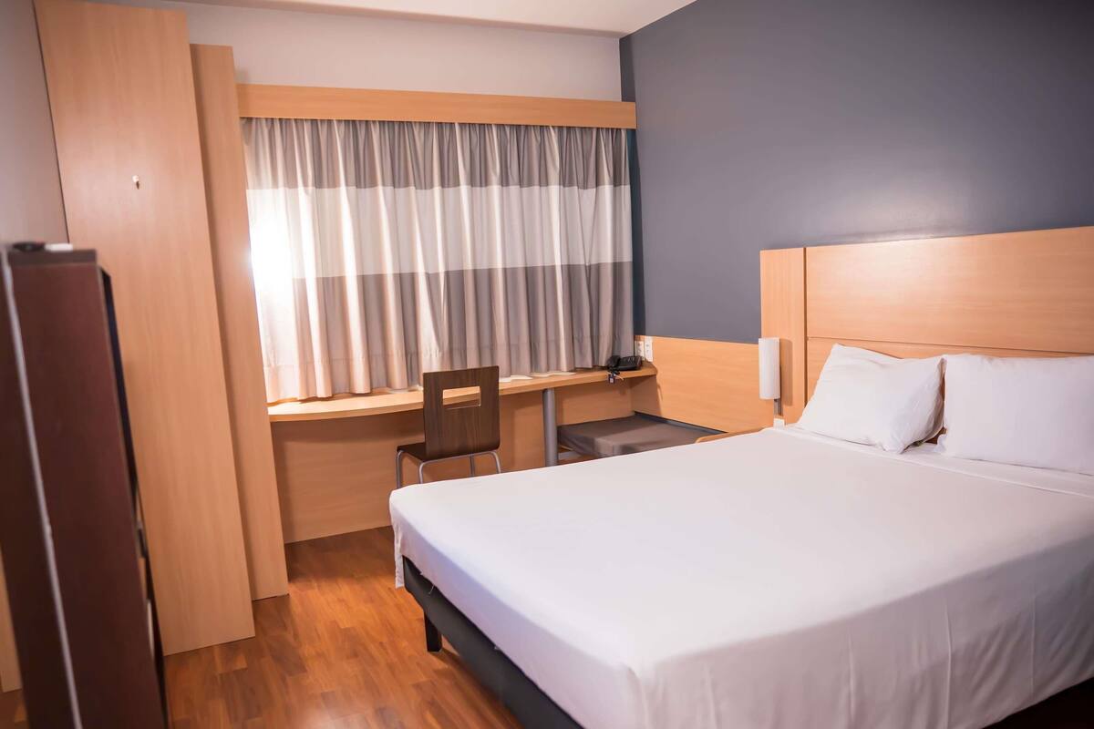 Ein Hotelzimmer mit Bett, Schreibtisch, Stuhl, einem Fenster mit Vorhängen und Holzboden.