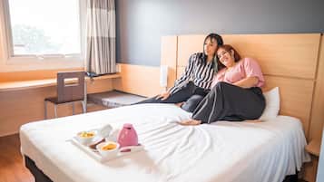 Habitación de hotel con cama, escritorio y silla. Cuenta con una ventana con cortinas, y una bandeja con comida y una botella de agua rosa sobre la cama.