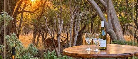 An outdoor dining area with a table and two stools, surrounded by greenery and trees.