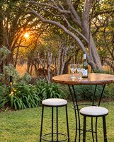 An outdoor dining area with a table and two stools, surrounded by greenery and trees.