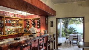 A bar area with wooden shelves stocked with bottles and glasses, a counter with stools, and a view of the outdoors through a glass door. - Musketeers Lodge (Bulawayo)