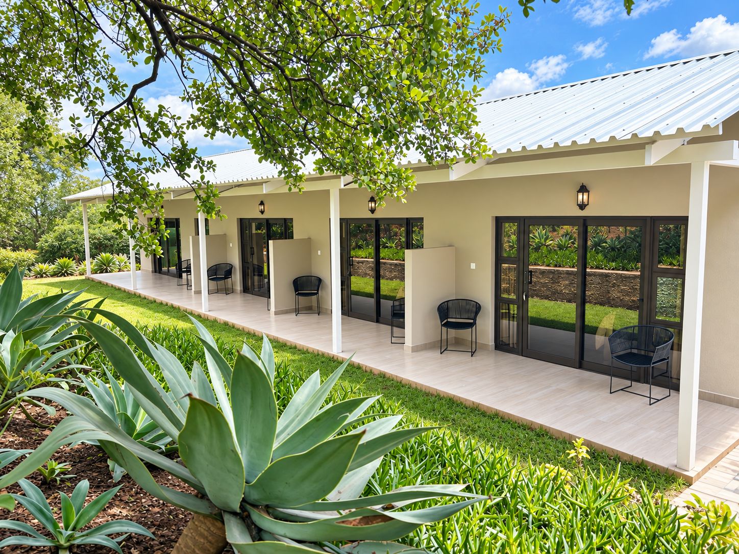 A covered outdoor area with chairs and a pathway, surrounded by greenery and a building with large windows.