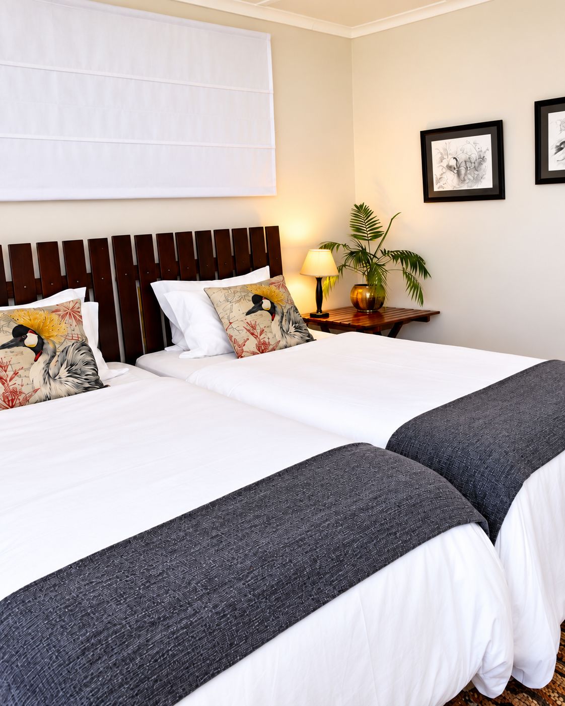 A hotel room with two beds, a nightstand with a lamp, a potted plant, and framed artwork on the wall.