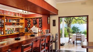 Zona de bar con estanterías de madera llenas de botellas y vasos, un barman y vistas a un jardín a través de un arco abierto.