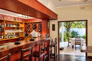 A bar area with wooden shelves stocked with bottles and glasses, a bartender, and a view of a garden through an open doorway. - Musketeers Lodge (Bulawayo)