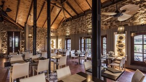 Un restaurant spacieux avec de hauts plafonds, des murs en pierre et de grandes fenêtres. Les tables sont disposées de manière informelle, et des lampes suspendues éclairent l’espace.