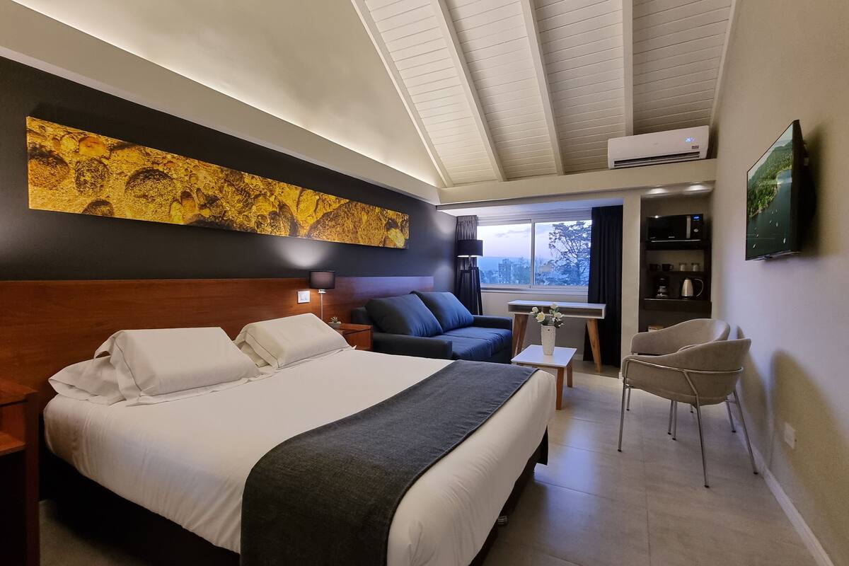 Ett modernt hotellrum med en stor säng, en soffa, ett litet bord och en stol. Det finns en tavla på väggen, ett fönster med utsikt och en luftkonditioneringsanläggning.