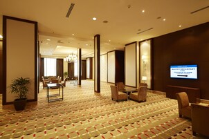 Un vestíbulo de hotel con alfombra estampada, zonas de estar, una pantalla de televisión mostrando información de eventos y una planta en maceta.